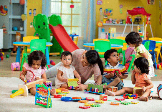 A teacher and five young children sitting on a rug in a brightly colored classroom surrounded by educational toys, blocks, and a small red slide.