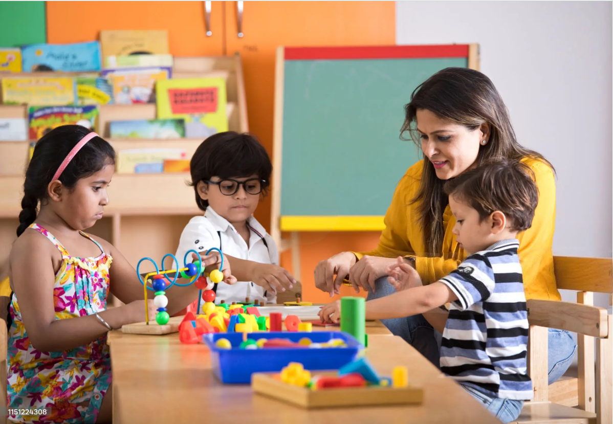 Smiling teacher helps three diverse students play with colored blocks in classroom.